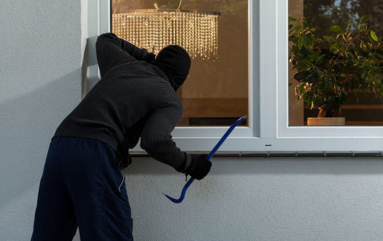 Einbrecher mit Skimaske und Brecheisen blickt durch ein Fenster ins Haus.
