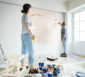 Ehepaar streicht gemeinsam die Wohnzimmerwand am Boden stehen viele Farbeimer und Malutensilien.
